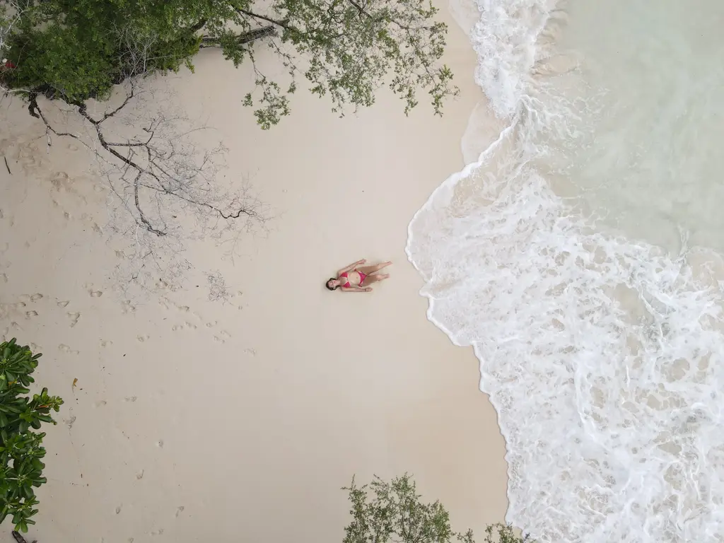 Aerial view looking straight down at a person lying on pale sand at the waterline, with white surf breaking beside them