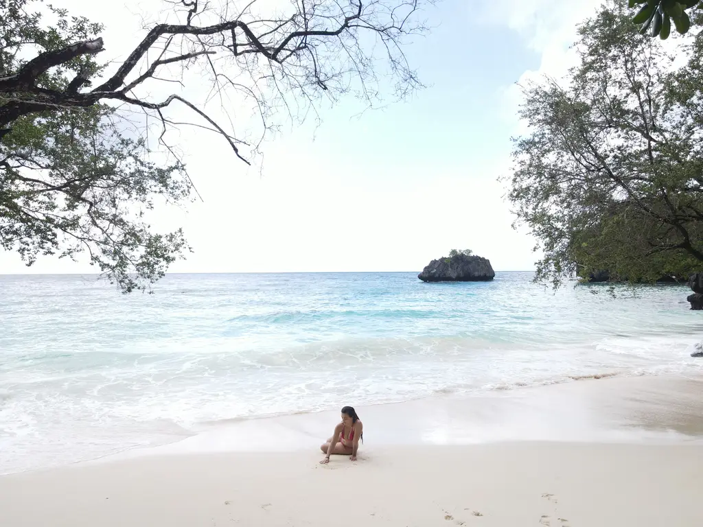 A person sitting in the shallows on a white sand beach, with a single rock standing offshore