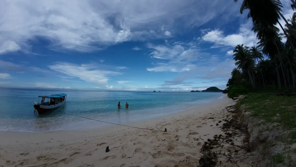 A wooden boat moored off a beach with two people standing in the shallows under a wide sky