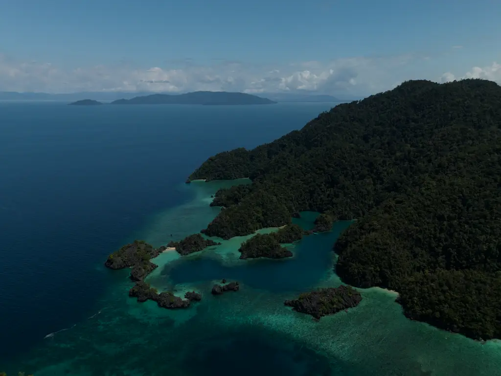 A forested headland dropping to shallow turquoise water, with small karst islets scattered offshore