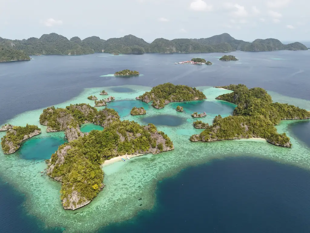 A wide cluster of green-topped limestone islands ringed by pale turquoise shallows, with deep blue water in front and a low mountain ridge on the horizon