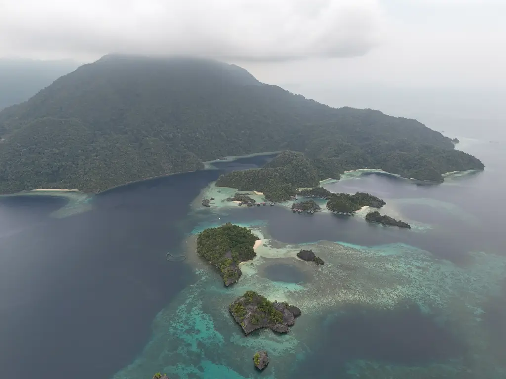 A wide bay in flat hazy light, low green islets over pale shallows with a long forested ridge behind