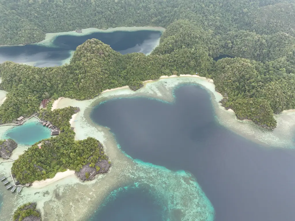 A deep blue lagoon enclosed by forested karst, with pale sand rims and buildings on stilts at the water edge