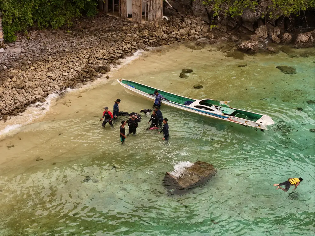 Passengers stepping from a wooden boat into shallow water beside a rocky shore, with no pier